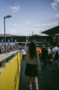 A woman takes a photo at a bustling outdoor sports event with a view of the stadium.