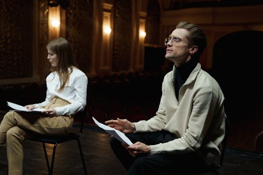 Two actors rehearsing with scripts in a dimly lit theater auditorium.