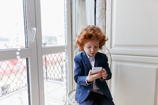 Red-haired boy wearing a suit using a smartphone indoors, seated near a window.