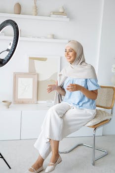 Smiling woman wearing hijab, recording a video indoors with a ring light and camera.